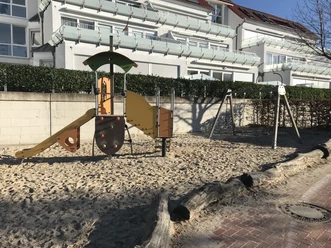 Ein moderner Spielplatz mit Sandboden, einer bunten Rutsche, Schaukel und Klettergerüst vor einer Ferienanlage.
