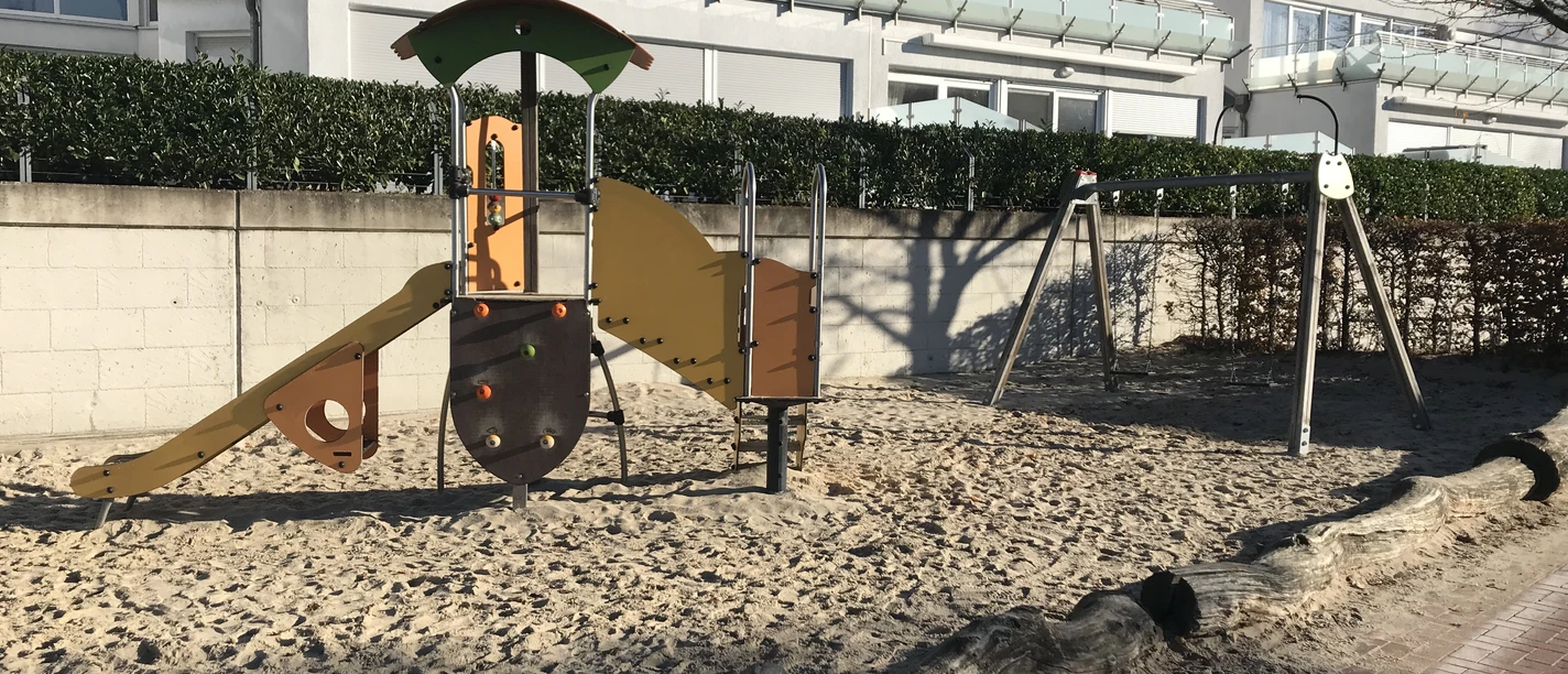 Ein moderner Spielplatz mit Sandboden, einer bunten Rutsche, Schaukel und Klettergerüst vor einer Ferienanlage.