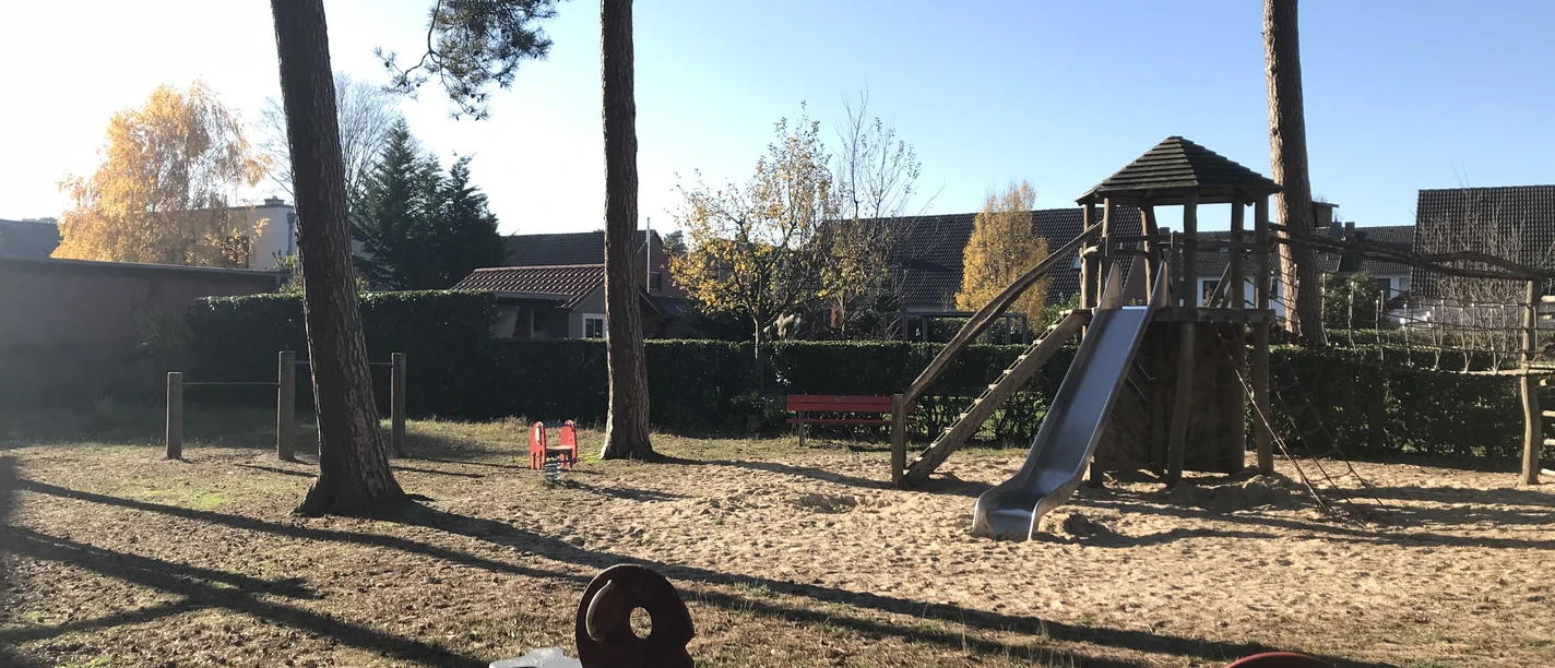 Spielplatz mit Kletterturm, Rutsche und Wipptieren unter hohen Bäumen im herbstlichen Sonnenlicht.