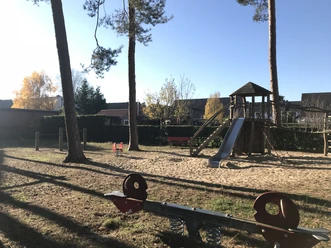 Spielplatz Schlesierweg Spielplatz mit Kletterturm, Rutsche und Wipptieren unter hohen Bäumen im herbstlichen Sonnenlicht.