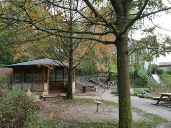 Ein Pavillon steht unter herbstlichen Bäumen, daneben ein geschlungener Metallspielplatz in einem Parksetting.