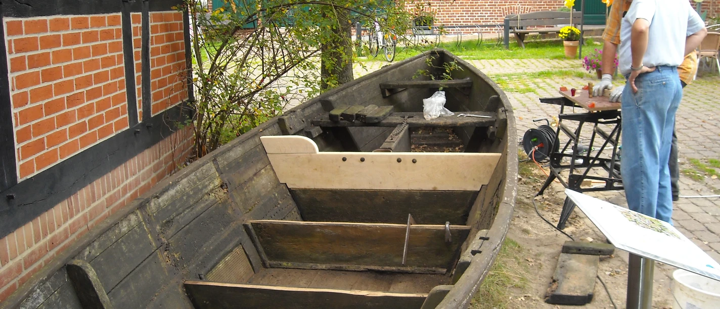 Das Bild zeigt ein altes, teilweise restauriertes Holzboot auf einem Platz vor einem Backsteingebäude.