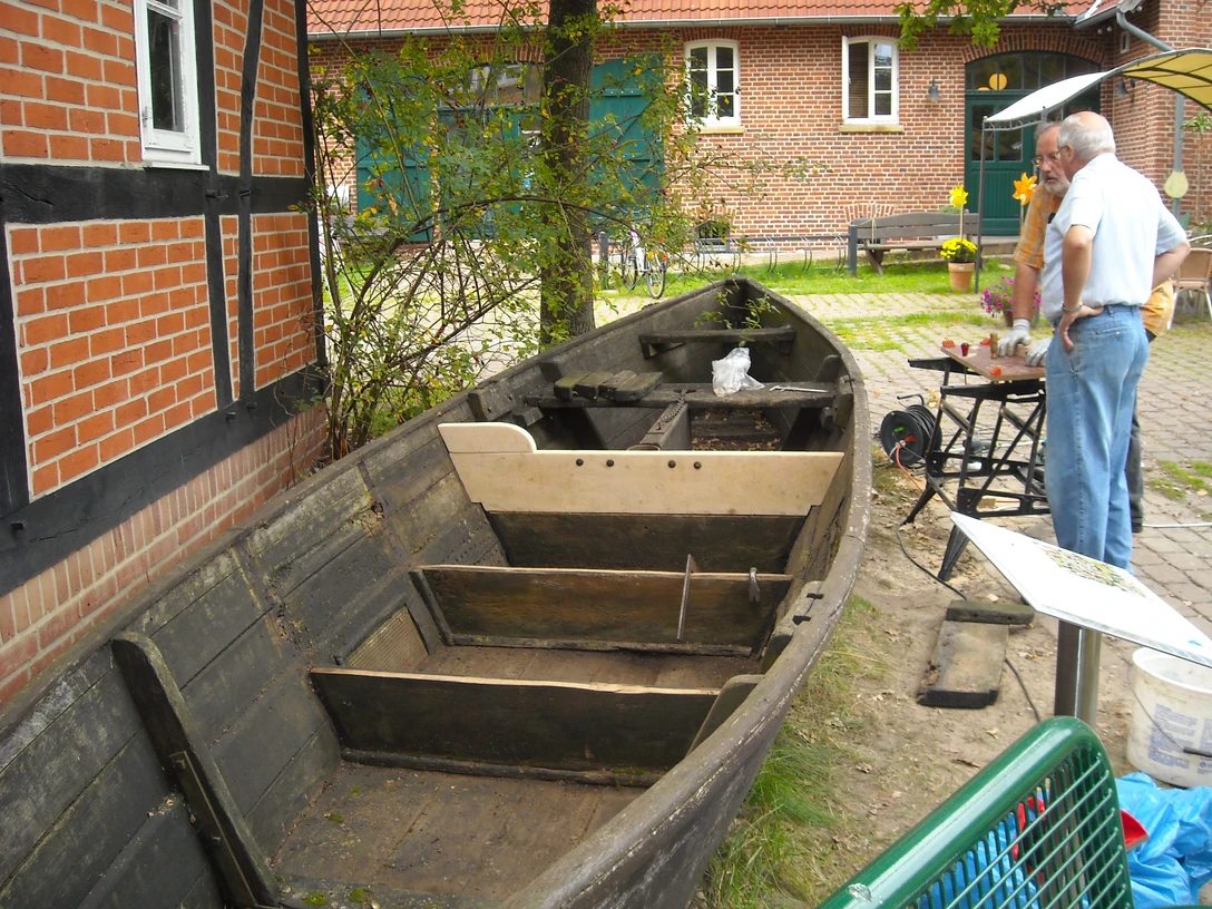 Restaurierung "Heske Deern" Das Bild zeigt ein altes, teilweise restauriertes Holzboot auf einem Platz vor einem Backsteingebäude.