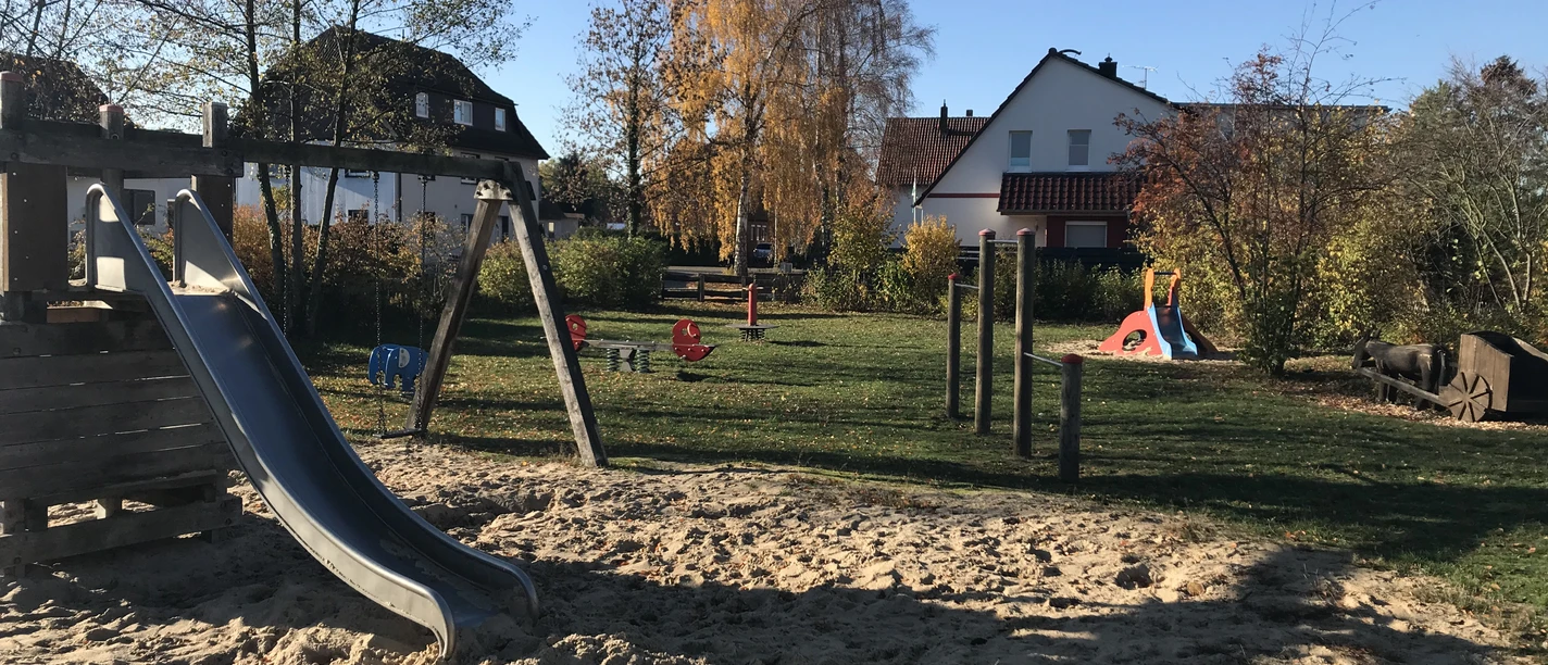 Ein moderner Spielplatz mit Klettergerüst, Rutsche und Wippe, eingerahmt von herbstlichen Bäumen.