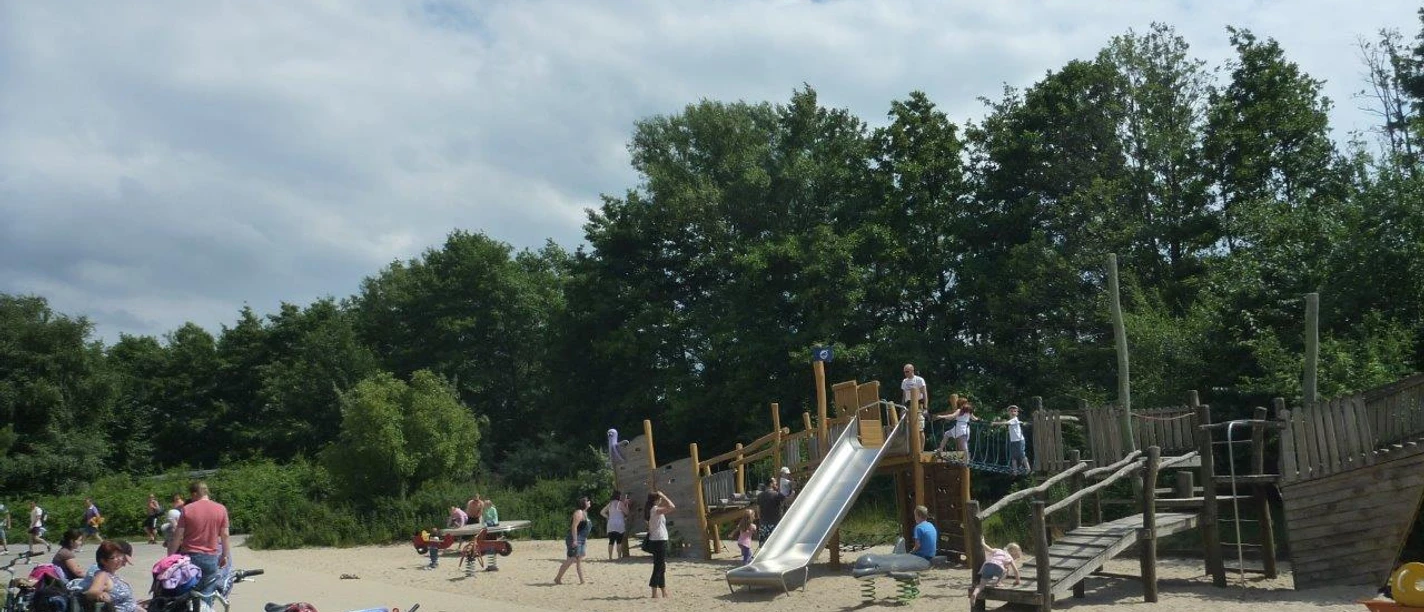 Spielplatz Badeinsel Ein lebendiger Spielplatz mit Holzkonstruktionen, Rutsche und Kindern beim Spielen im sonnigen Park.