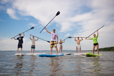 Surfers Paradise Gruppe von sechs Menschen beim Stand-Up-Paddeln auf einem ruhigen See, Sonne und blauer Himmel im Hintergrund.