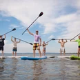 Surfers Paradise Gruppe von sechs Menschen beim Stand-Up-Paddeln auf einem ruhigen See, Sonne und blauer Himmel im Hintergrund.