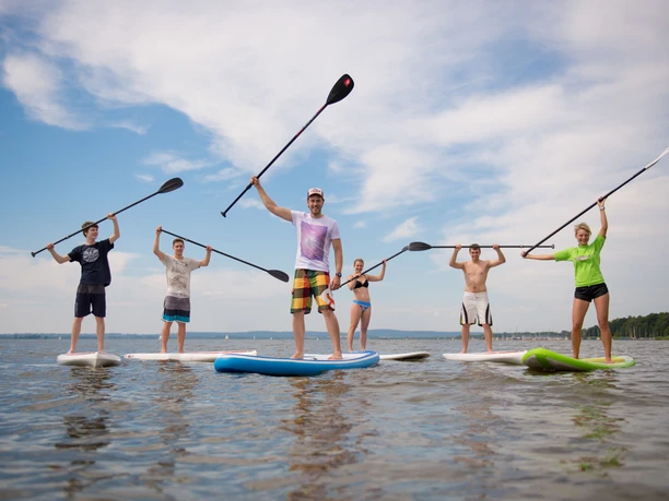 Surfers Paradise Gruppe von sechs Menschen beim Stand-Up-Paddeln auf einem ruhigen See, Sonne und blauer Himmel im Hintergrund.