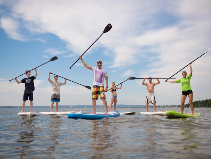 Surfers Paradise Gruppe von sechs Menschen beim Stand-Up-Paddeln auf einem ruhigen See, Sonne und blauer Himmel im Hintergrund.