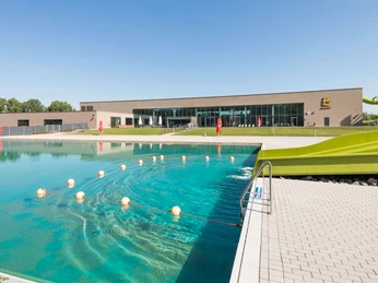balneon1-schwimmen-baden Ein modernes Freibad mit einem großen Schwimmerbecken neben einer Rutsche, umgeben von Grünflächen.