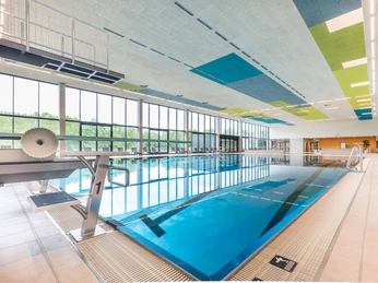 balneon3-schwimmen-baden Innenansicht eines modernen Schwimmbads mit Sprungbrettern und großen Fenstern, die Tageslicht einlassen.