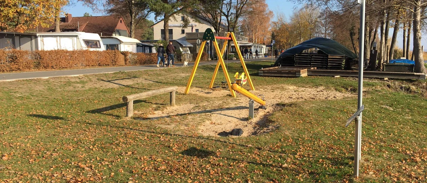 Spielplatz mit Schaukel und Sandkasten auf einer sonnigen Wiese, umrahmt von Bäumen und Wohnhäusern.