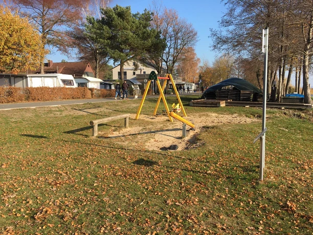 A playground with a swing and a sandbox on a sunny meadow, surrounded by trees and houses.