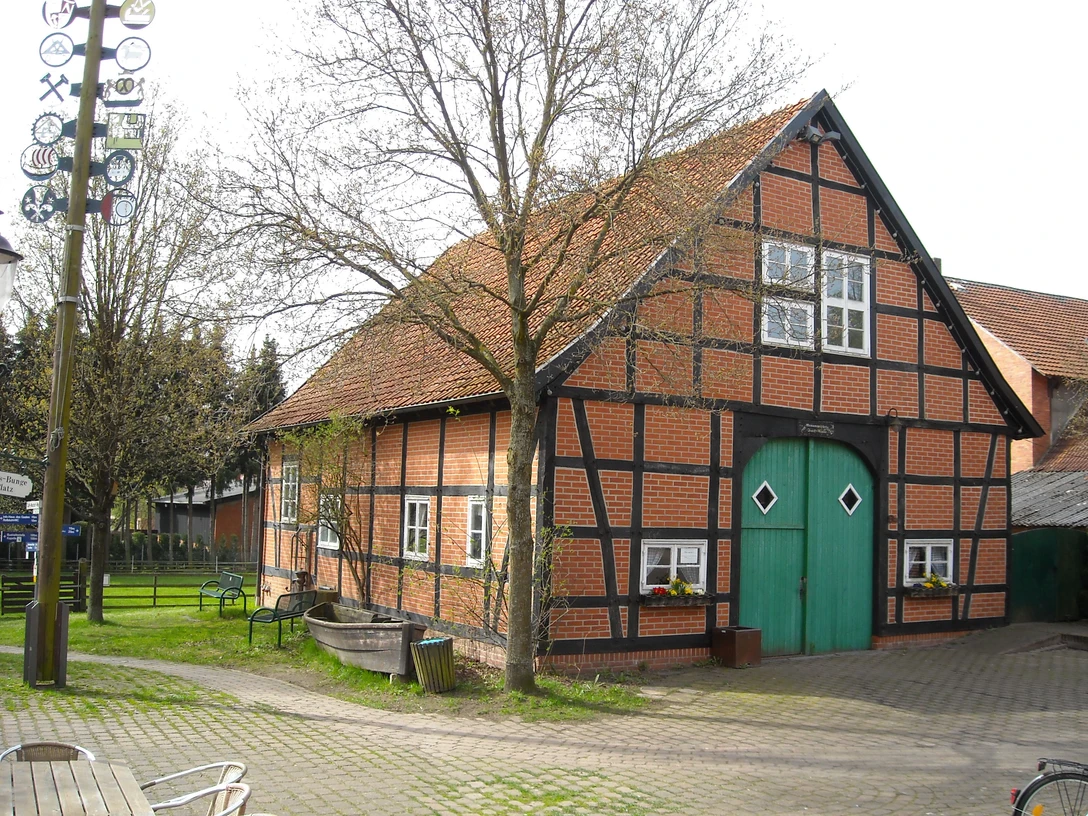 Fachwerkhof im Grünen, mit rotem Backstein und blauem Tor; umgeben von Bäumen und Bänken.