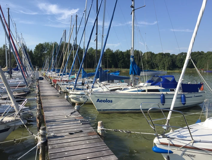 Steggemeinschaft Steg N17 + N18 - GbR Ein idyllischer Holzsteg am See mit zahlreichen, farbenfrohen Segelbooten, gesäumt von Bäumen.