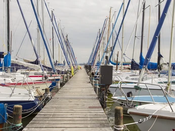 Boote am Steg Reihe von Segelbooten am hölzernen Steg unter bewölktem Himmel, umgeben von ruhigem Wasser.