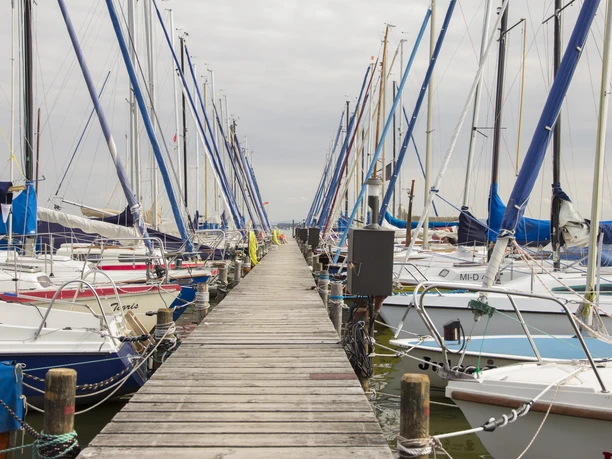 Boote am Steg Reihe von Segelbooten am hölzernen Steg unter bewölktem Himmel, umgeben von ruhigem Wasser.