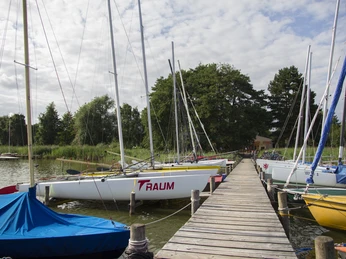 Steggemeinschaft Steg N17 + N18 mit Blick auf den Uferweg Auf einem Holzsteg sind Segelboote vertäut, flankiert von üppigem Grün und einem klaren Himmel. Ein friedlicher Anblick am Uferweg.
