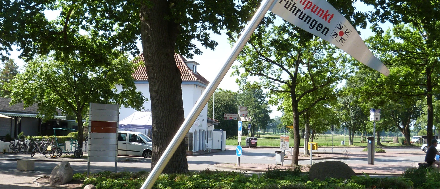 Treffpunkf für Gästeführungen Eine Fahne mit der Aufschrift "Treffpunkt Führungen" neigt sich neben einem Weg in einem grünen Parkbereich.