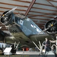 In der Ju 52 Halle am Fliegerhorst Wunstorf steht eine historische Junkers Ju 52. Das Flugzeug mit seinen ikonischen drei Motoren ist in einer Halle mit hölzernen Balken untergebracht. Zwei Figuren in historischen Uniformen stehen daneben.