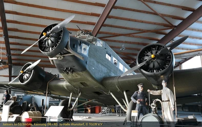 Ju 52 Halle am Fliegerhorst Wunstorf In der Ju 52 Halle am Fliegerhorst Wunstorf steht eine historische Junkers Ju 52. Das Flugzeug mit seinen ikonischen drei Motoren ist in einer Halle mit hölzernen Balken untergebracht. Zwei Figuren in historischen Uniformen stehen daneben.