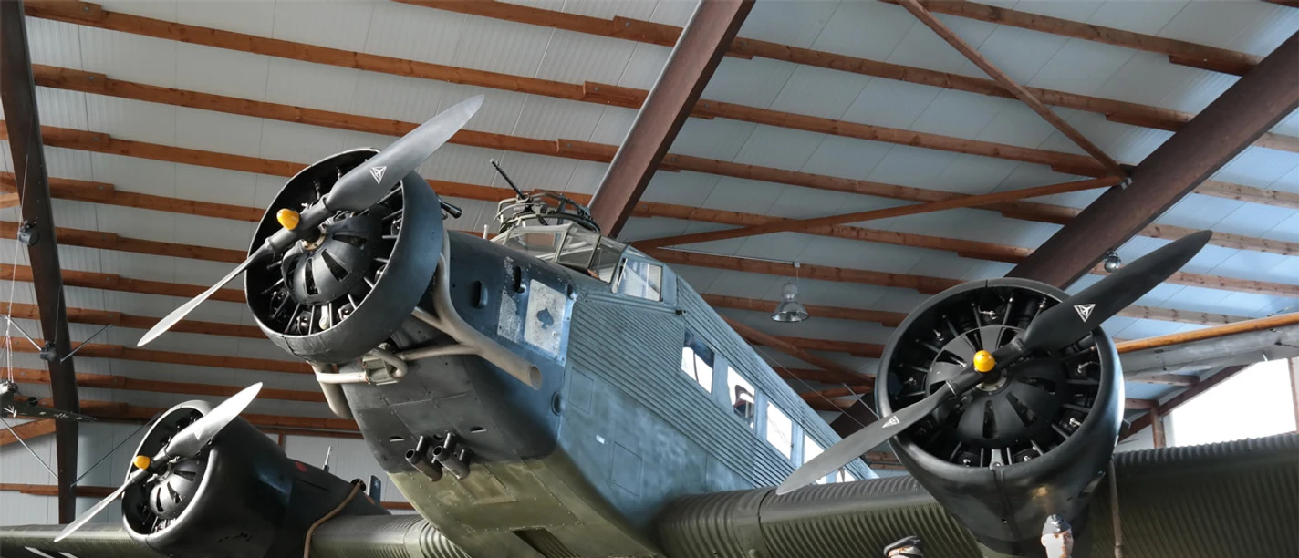 Ju 52 Halle am Fliegerhorst Wunstorf In der Ju 52 Halle am Fliegerhorst Wunstorf steht eine historische Junkers Ju 52. Das Flugzeug mit seinen ikonischen drei Motoren ist in einer Halle mit hölzernen Balken untergebracht. Zwei Figuren in historischen Uniformen stehen daneben.