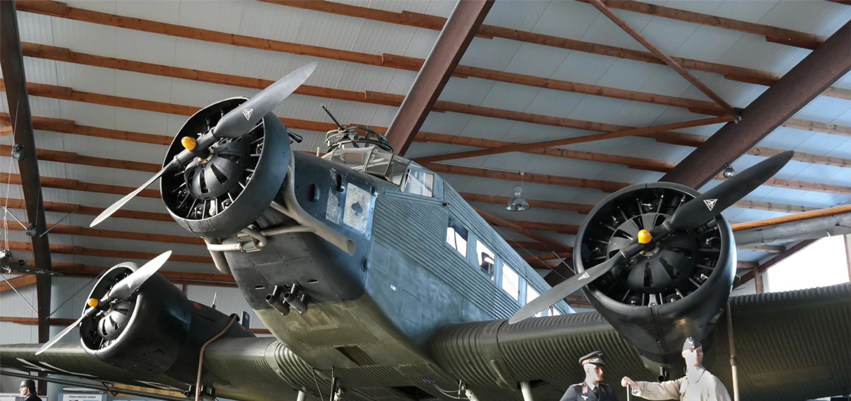 Ju 52 Halle am Fliegerhorst Wunstorf In der Ju 52 Halle am Fliegerhorst Wunstorf steht eine historische Junkers Ju 52. Das Flugzeug mit seinen ikonischen drei Motoren ist in einer Halle mit hölzernen Balken untergebracht. Zwei Figuren in historischen Uniformen stehen daneben.A historic Junkers Ju 52 stands in the Ju 52 hangar at Wunstorf Air Base. The aircraft with its iconic three engines is housed in a hangar with wooden beams. Two figures in historical uniforms stand next to it.En historisk Junkers Ju 52 står i Ju 52-hangaren på Wunstorf Air Base. Flyet med de ikoniske tre motorer er anbragt i en hangar med træbjælker. To personer i historiske uniformer står ved siden af det.Een historische Junkers Ju 52 staat in de Ju 52 hangar op vliegbasis Wunstorf. Het vliegtuig met zijn iconische drie motoren staat in een hangar met houten balken. Twee figuren in historische uniformen staan ernaast.