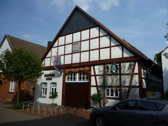 Steinhuder Museen De foto toont een traditioneel vakwerkhuis met een steile gevel en houten decoraties, dat deel uitmaakt van het Steinhuder Museum. Fietsenrekken en een boom zijn zichtbaar voor het gebouw, terwijl rechts een geparkeerde auto te zien is. De lucht is licht bewolkt.