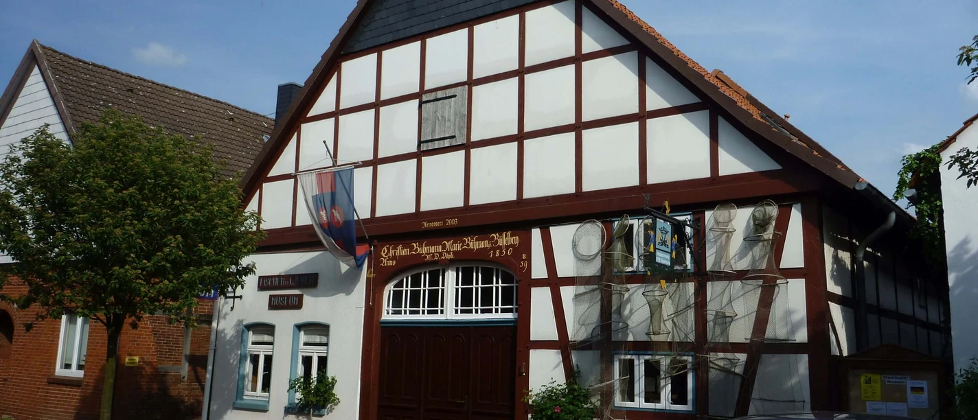Steinhuder Museen The picture shows a traditional half-timbered house with a steep gable and wooden decorations, which is part of the Steinhuder Museum. Bicycle stands and a tree are visible in front of the building, while a parked car can be seen on the right. The sky is slightly cloudy.