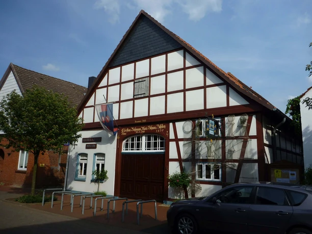 Steinhuder Museen Das Bild zeigt ein traditionelles Fachwerkhaus mit einem steilen Giebel und hölzernen Verzierungen, das Teil des Steinhuder Museums ist. Vor dem Gebäude sind Fahrradständer und ein Baum sichtbar, während rechts ein geparktes Auto zu sehen ist. Der Himmel ist leicht bewölkt.