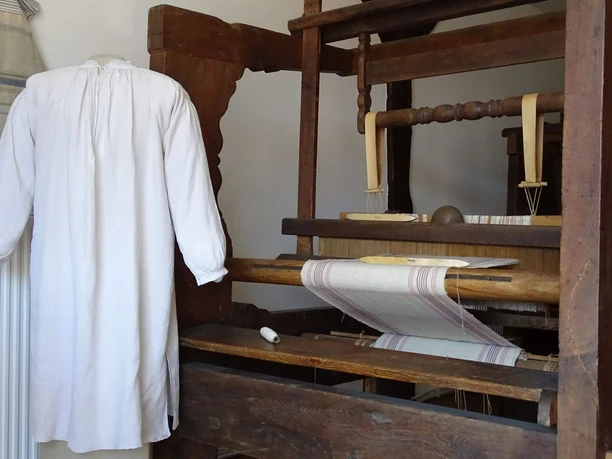 Hemd ohne Naht A plain white long-sleeved shirt hangs next to a historic wooden loom on which light-colored lengths of fabric are woven.