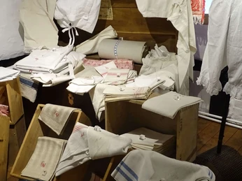 Aussteuertruhe Historische Aussteuertruhe umgeben von schweren weißen Leinen, fein gestickten Handtüchern und alten Betttüchern, die sich in einem Ausstellungsraum befinden.Historic dowry chest surrounded by heavy white linen, finely embroidered towels and old bed sheets in an exhibition room.Historisk medgiftskiste omgivet af tungt hvidt linned, fint broderede håndklæder og gamle sengelinned i et udstillingsrum.Historische bruidskist omringd door zwaar wit linnen, fijn geborduurde handdoeken en oude lakens in een tentoonstellingsruimte.