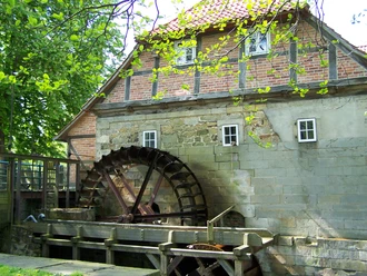 Historische Wassermühle Laderholz mit rotem Ziegeldach und großem Holzrad in grüner Umgebung.