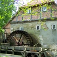 Historische Wassermühle Laderholz mit rotem Ziegeldach und großem Holzrad in grüner Umgebung.