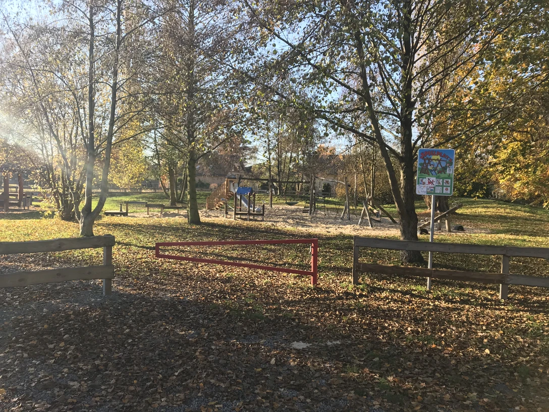Spielplatz in herbstlicher Landschaft mit Schaukeln und Rutsche, umgeben von Bäumen und Grünfläche.