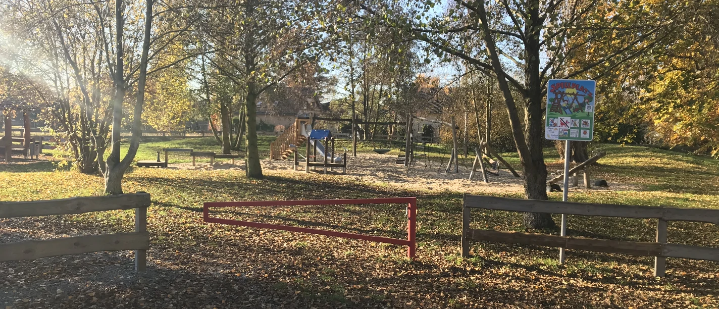 Spielplatz Försterwiese Spielplatz in herbstlicher Landschaft mit Schaukeln und Rutsche, umgeben von Bäumen und Grünfläche.