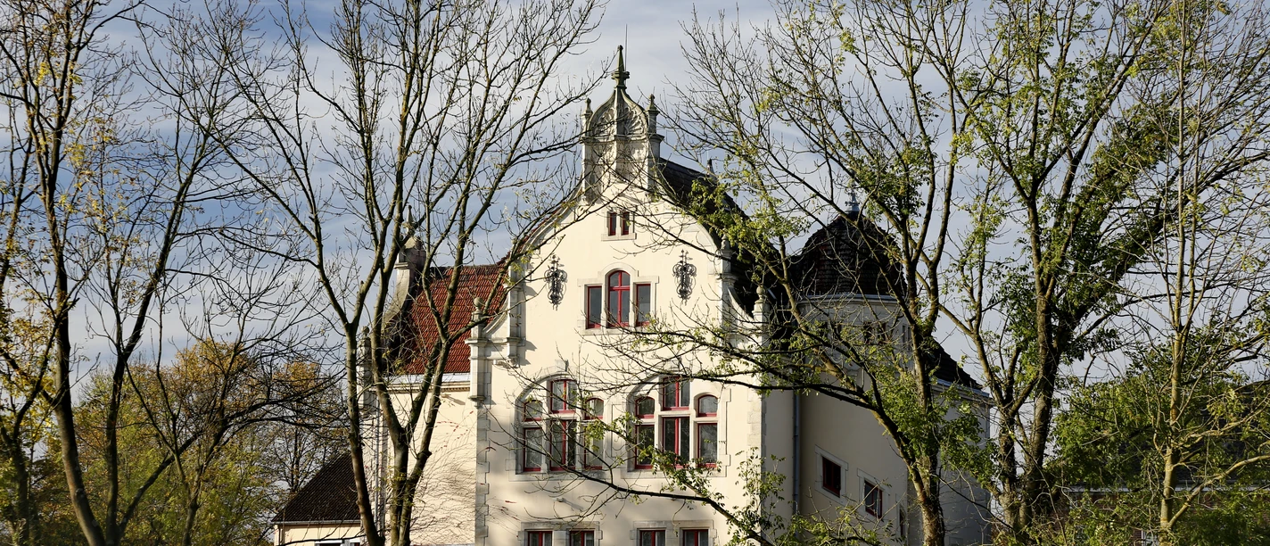 Ein historisches Gebäude mit verzierten Fenstern und Turmspitze, umrahmt von Bäumen im Herbst.