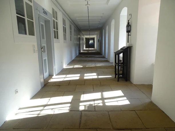 Long, white monastery corridor with thick stone slabs, windows and points of light on the floor.