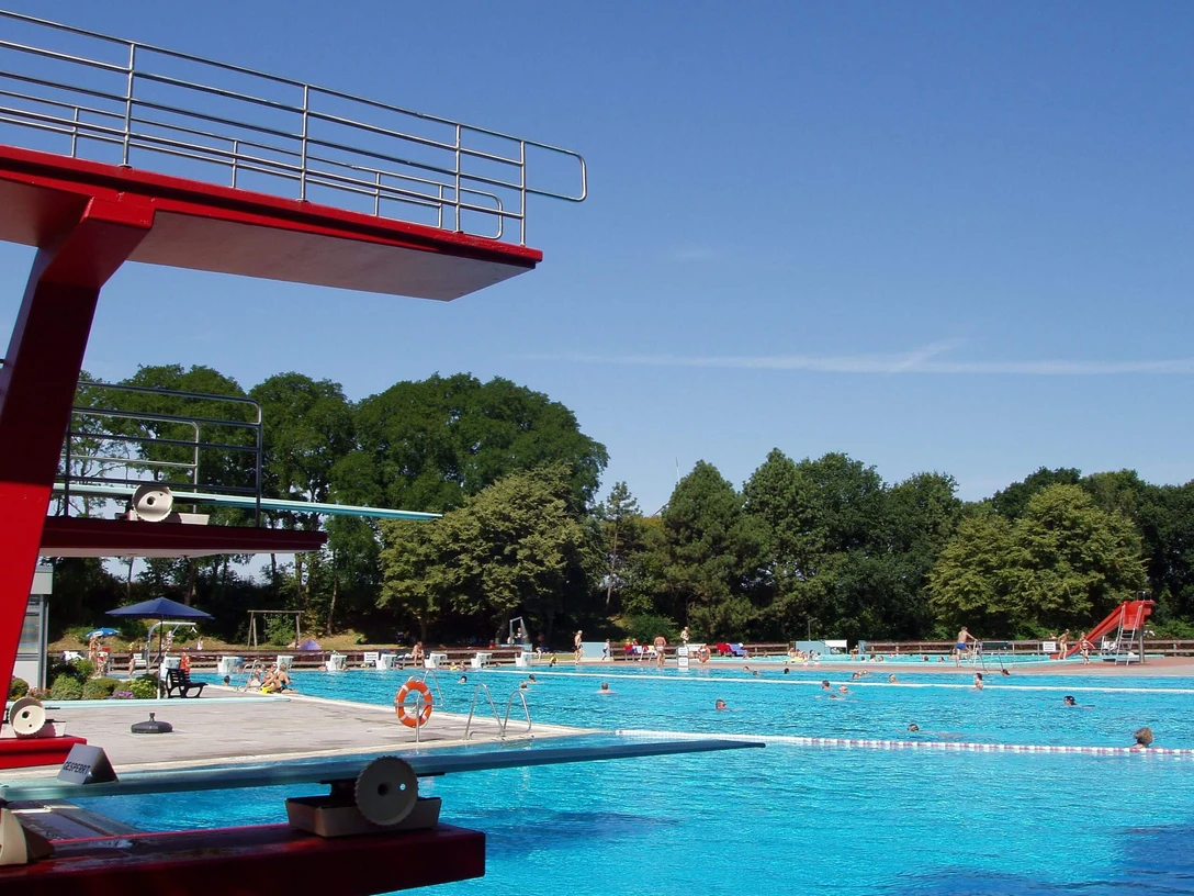 Freibad Bokeloh Großes Freibad mit Sprungturm und Becken, umgeben von Bäumen und sommerlich blauem Himmel.