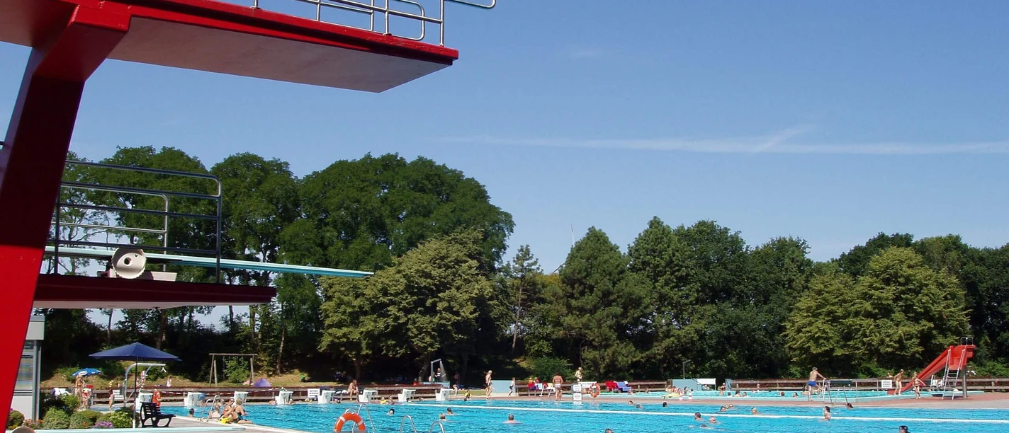 Freibad Bokeloh Großes Freibad mit Sprungturm und Becken, umgeben von Bäumen und sommerlich blauem Himmel.
