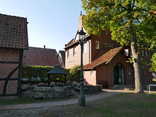 Historischer Backsteinbau mit markantem Schornstein, überdachtem Sitzbereich und umgebenden Bäumen.