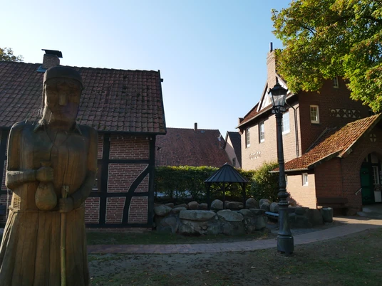Holzskulptur vor Fachwerkhäusern auf dem Aloys-Bunge-Platz, umgeben von Bäumen und einem Grillplatz.