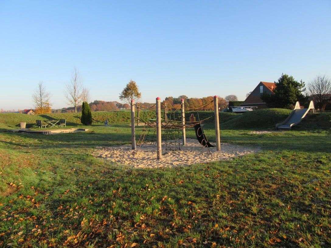 Spielplatz mit Klettergerüst auf Sand, grüner Wiese und Rutsche, umgeben von herbstlichen Bäumen.