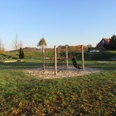 Spielplatz mit Klettergerüst auf Sand, grüner Wiese und Rutsche, umgeben von herbstlichen Bäumen.