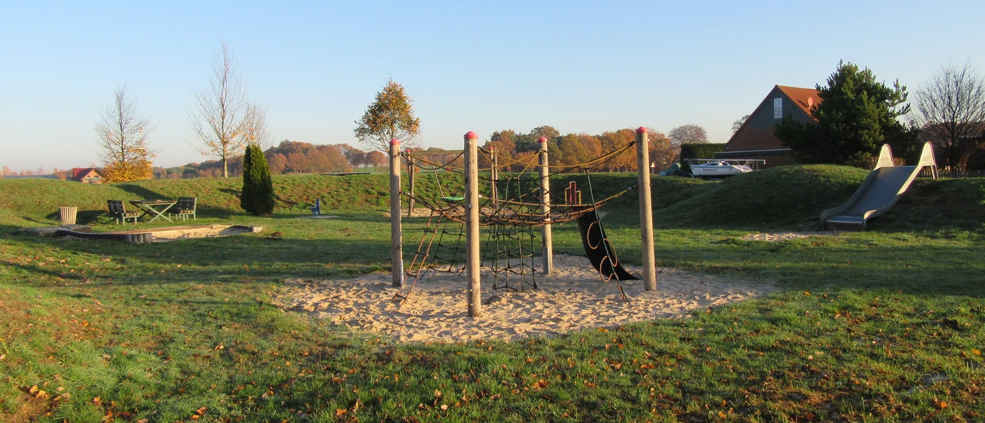 Spielplatz mit Klettergerüst auf Sand, grüner Wiese und Rutsche, umgeben von herbstlichen Bäumen.
