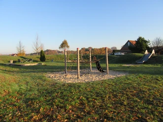 Foto Spielplatz Jägerstraße Spielplatz mit Klettergerüst auf Sand, grüner Wiese und Rutsche, umgeben von herbstlichen Bäumen.