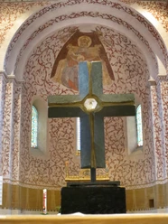 mandelsloh-st.osdag-kirche-kreuz-altar Ein großes Steinkreuz auf einem Sockel im Vordergrund, dahinter verzierte Wand mit gemalter Engelsfigur.