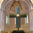 mandelsloh-st.osdag-kirche-kreuz-altar Ein großes Steinkreuz auf einem Sockel im Vordergrund, dahinter verzierte Wand mit gemalter Engelsfigur.