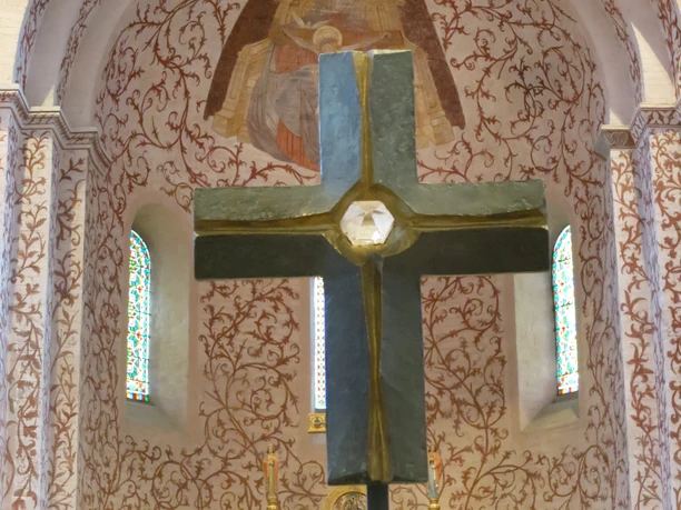 mandelsloh-st.osdag-kirche-kreuz-altar Ein großes Steinkreuz auf einem Sockel im Vordergrund, dahinter verzierte Wand mit gemalter Engelsfigur.