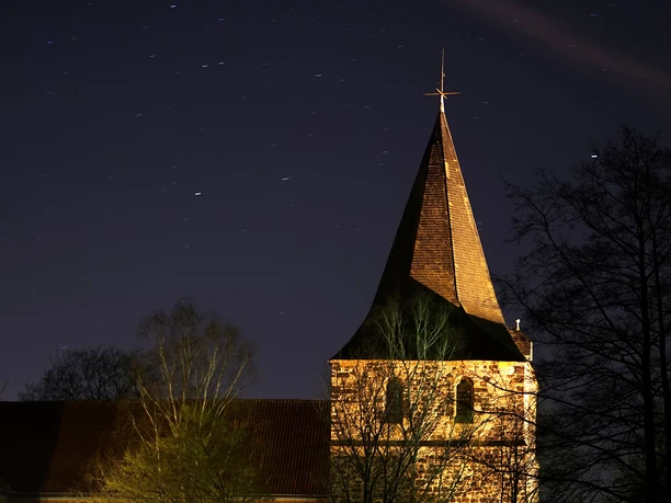 mandelsloh-st.osdag-kirche-nacht St. Osdag Kirche in Mandelsloh bei Nacht, beleuchtet unter klarem Sternenhimmel und umgeben von Bäumen.